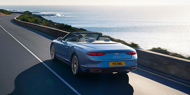 Bentley Hannover New Bentley Continental GTC Azure convertible rear view in Light Windsor blue paint with 22 inch ten spoke wheels driving dynamically on an ocean road in the sunshine